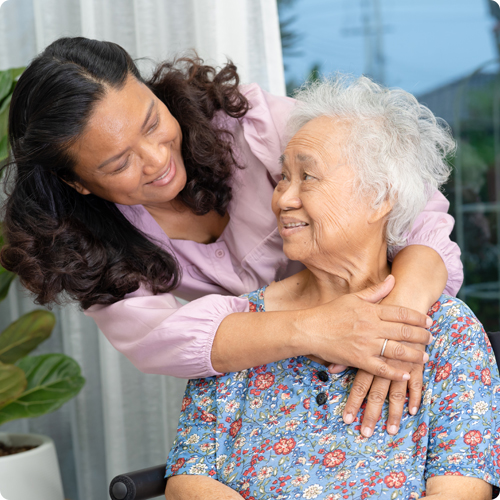 A middle-aged Asian woman smiling and leaning down to hug her elderly mother, who is seated in a wheelchair and looking back at her with affection.