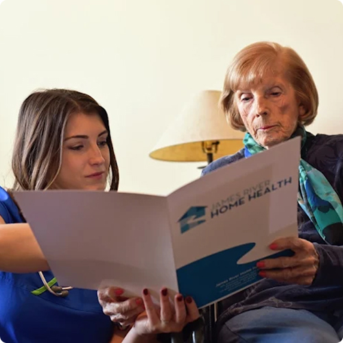 A friendly caregiver with long dark hair holds up an open James River Home Health services brochure, reviewing the information with an elderly woman seated next to her
