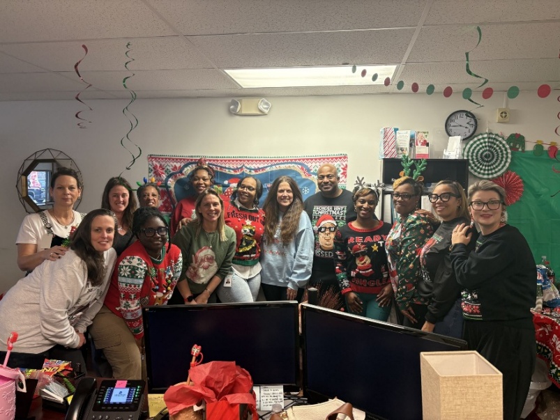 A group of thirteen diverse office colleagues smiling and posing together while wearing festive Christmas sweaters and holiday accessories in a decorated office setting.